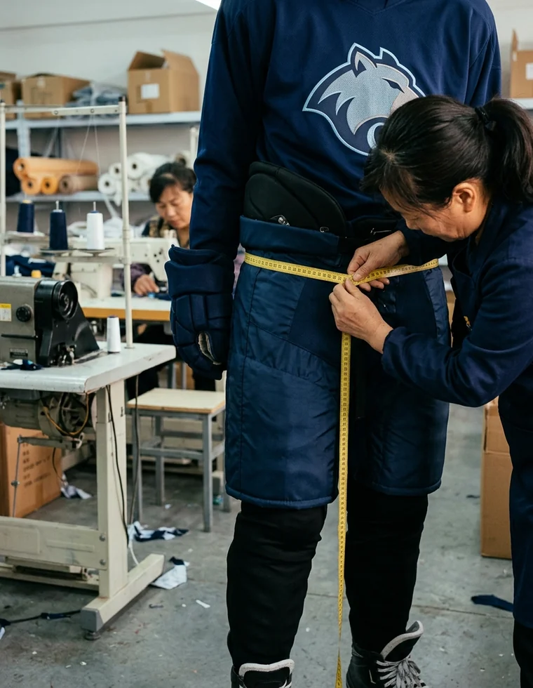 A tailor's tape measuring a hockey pant shell worn over a player's own padded pants, waist measurement taken in the dressed state