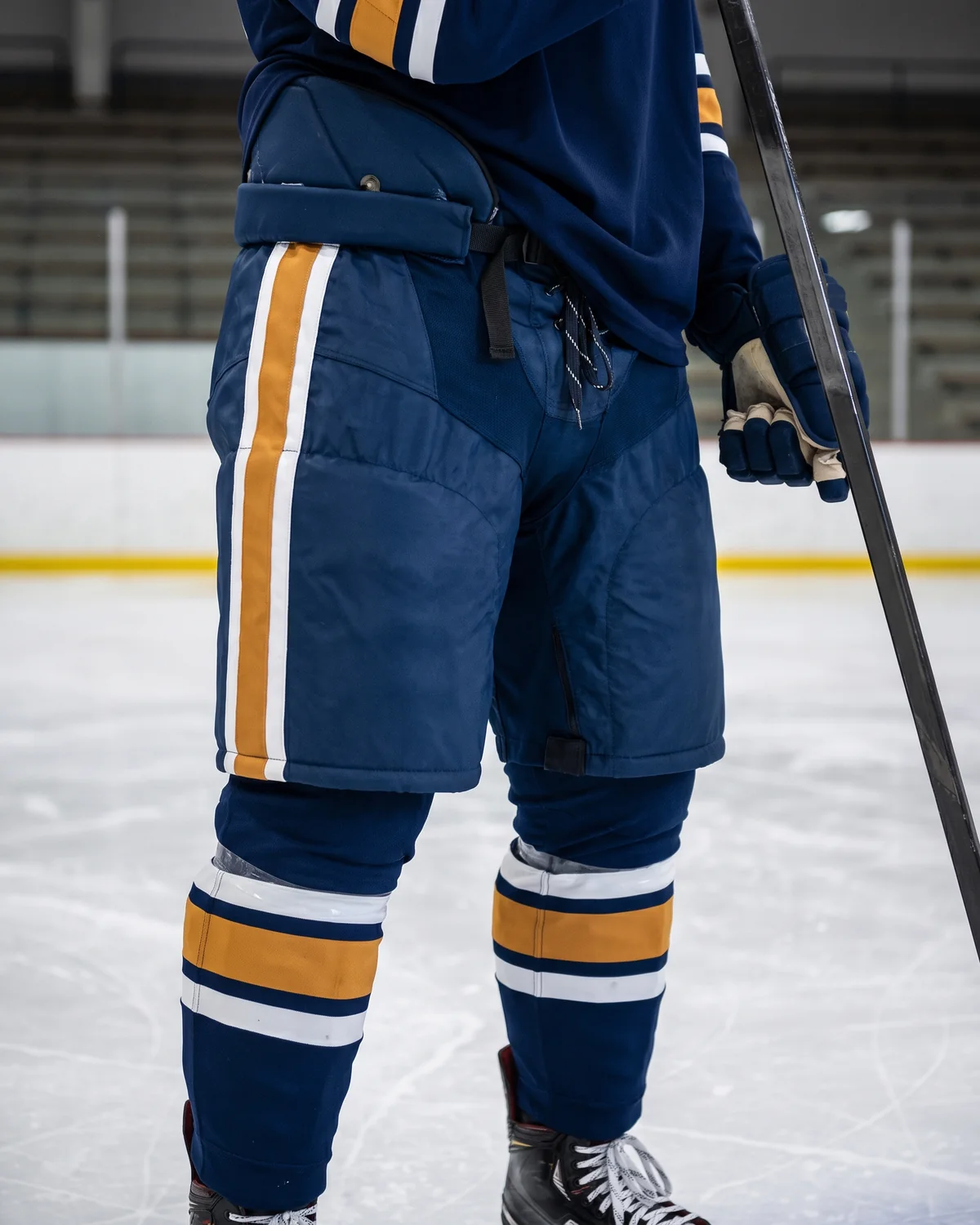 Illustrative image of a hockey player on the ice, seen from waist to skates, in a loose navy pant shell with a white and amber side stripe worn over padded hockey pants, with a drawstring waist, wide leg openings and matching striped socks