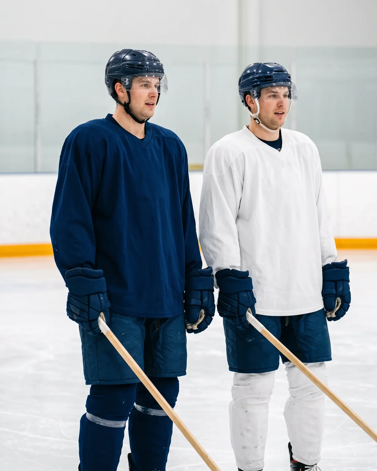 Two hockey players at an indoor rink practice wearing the same loose-fit practice jersey in two colorways, one solid navy and one solid white, with wide sleeves and a V-neck collar.